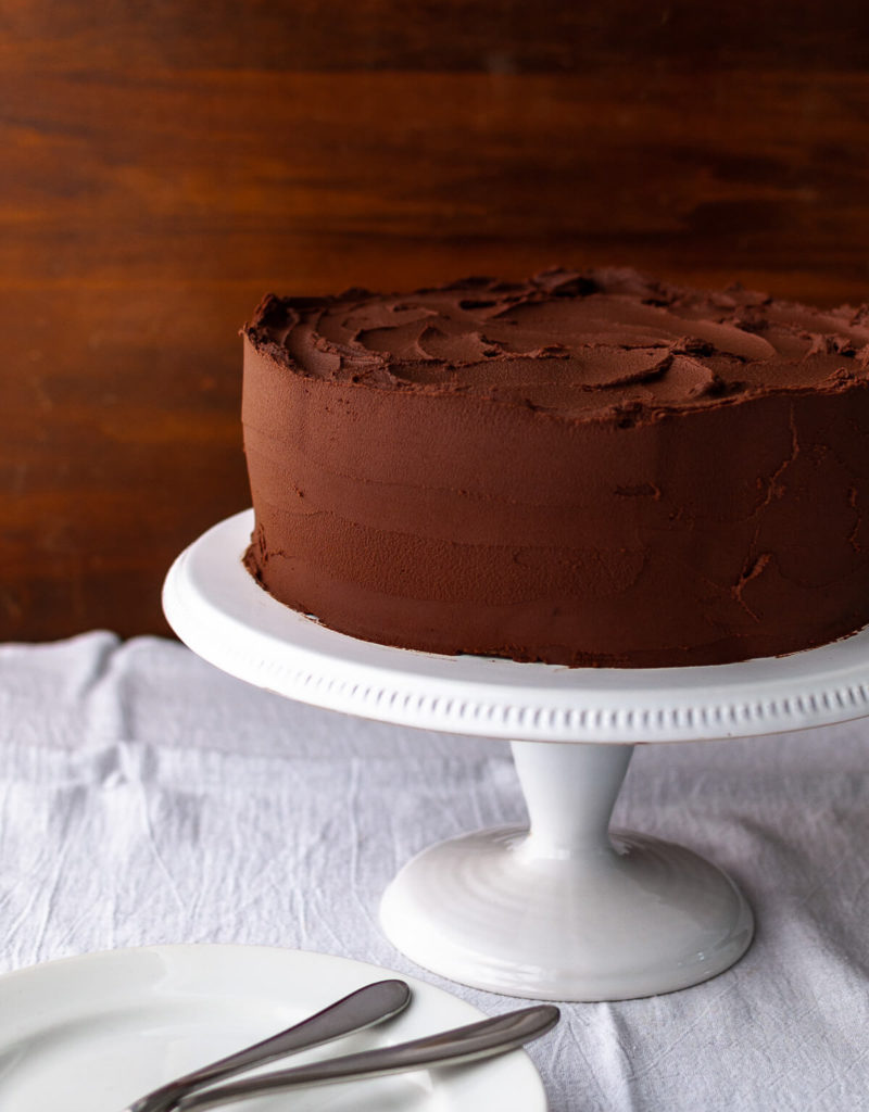 A chocolate cake with chocolate frosting on a white cake stand.