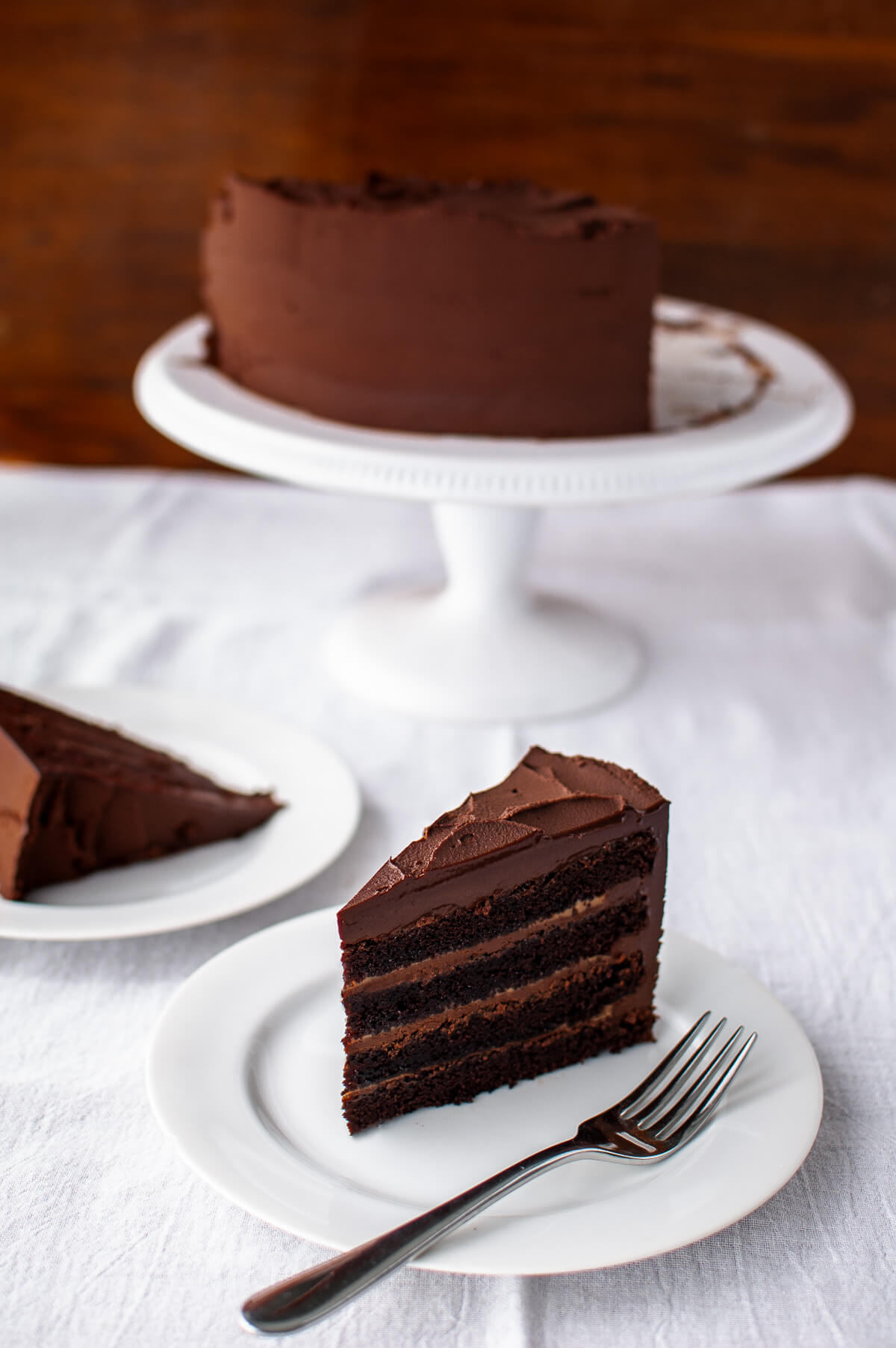 A slice of chocolate cake with caramel filling on a white plate next to a cake on a cake stand.