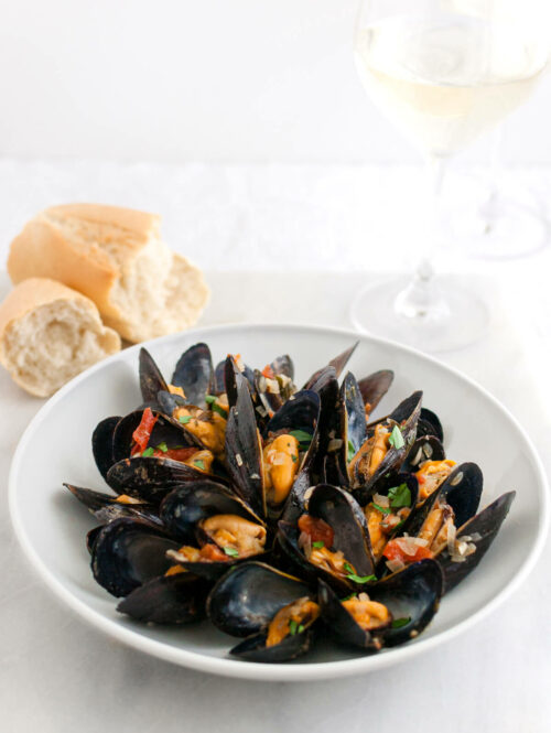 Cooked mussels in a white a bowl next to some french bread.