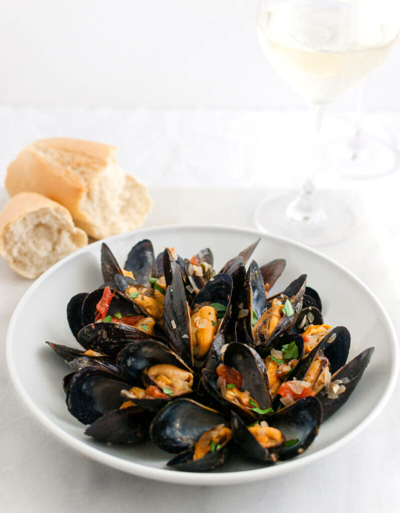 Cooked mussels in a white a bowl next to some french bread.