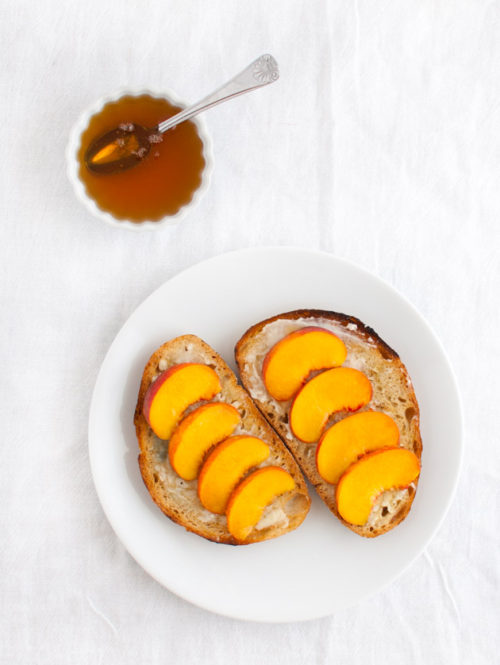 Peach slices on toasted bread on a white plate next to a bowl of honey.