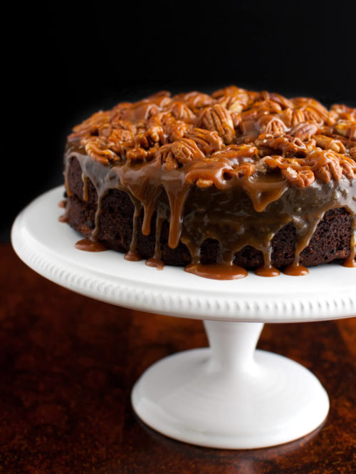 A chocolate cake topped with caramel sauce and toasted pecans on a white cake stand.