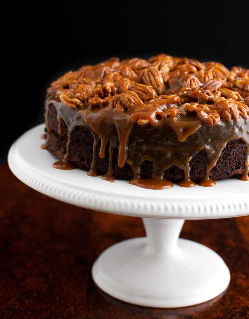 A chocolate cake topped with caramel sauce and toasted pecans on a white cake stand.