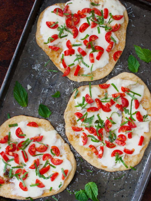 Three small pizzas topped with cheese and fresh basil and tomatoes on a metal tray.