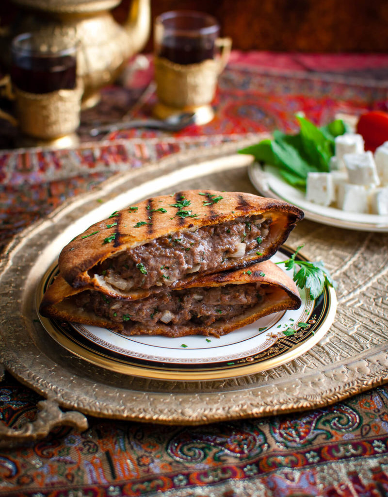 Grilled lamb burger in a pita on a china plate next to a plate of feta and fresh herbs.