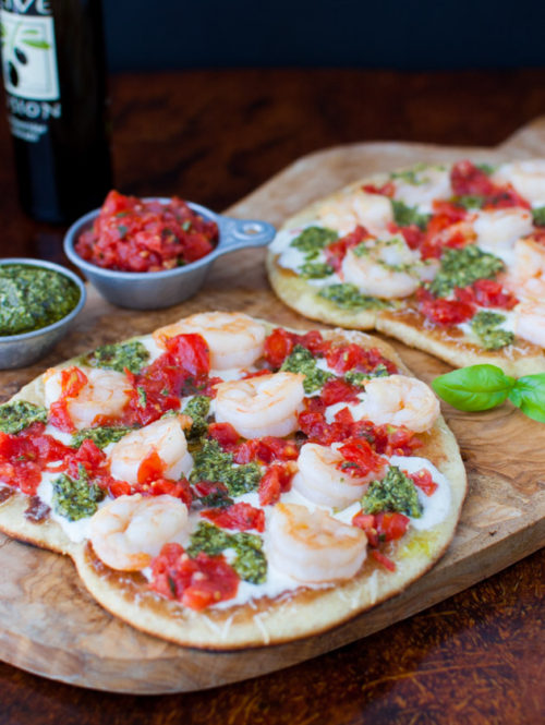 Shrimp pizzas on a wooden cutting board next to bowls of pesto and tomato sauce.