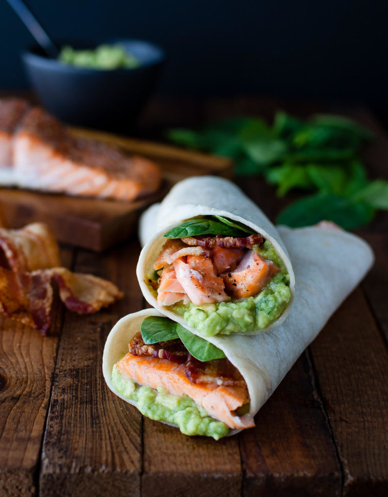Salmon, guacamole, bacon, and spinach wrapped inside a tortilla on a wooden board.