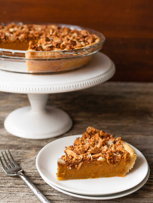 A slice of pumpkin pie on a plate next to a pie on a white cake stand.