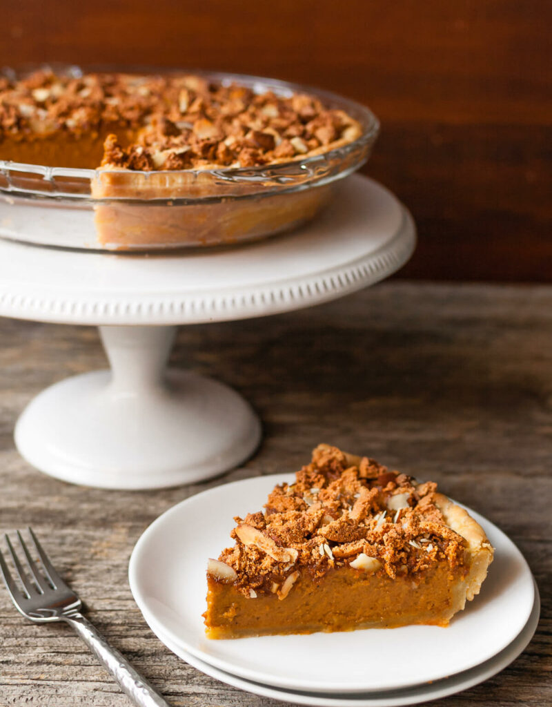 A slice of pumpkin pie on a plate next to a pie on a white cake stand.