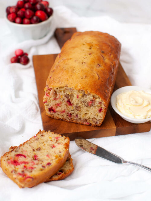 Loaf of cranberry bread on wooden board next to slices of bread and dish of softened butter.
