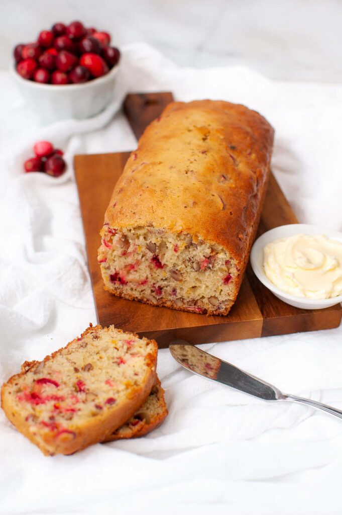 Loaf of cranberry bread on wooden board next to slices of bread and dish of softened butter.