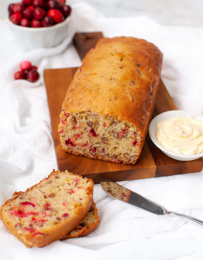 Loaf of cranberry bread on wooden board next to slices of bread and dish of softened butter.