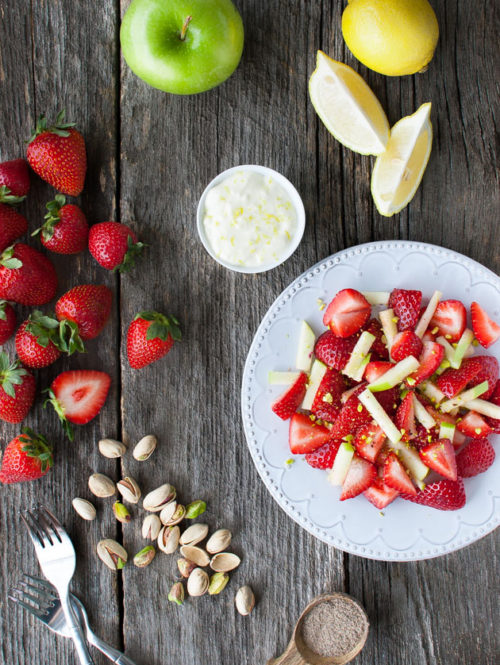 Fruit salad on a plate next to a green apple, lemon wedges, strawberries, and pistachios.