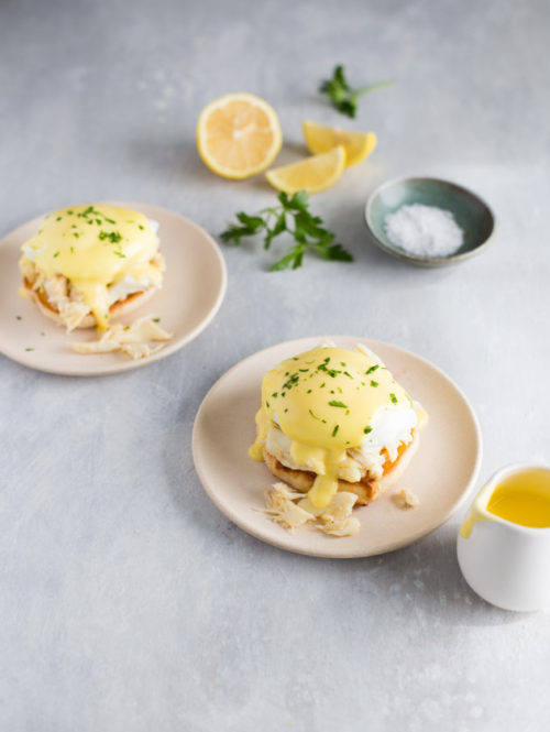Two small plates of crab eggs benedict next to a small pitcher of hollandaise sauce.