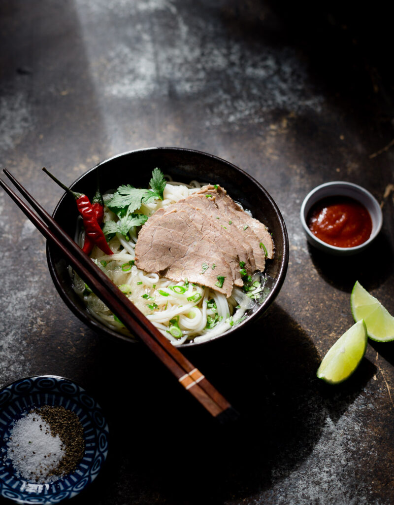 A bowl of lemongrass beef and noodle soup served with some hot sauce, lime wedges, and salt and pepper.
