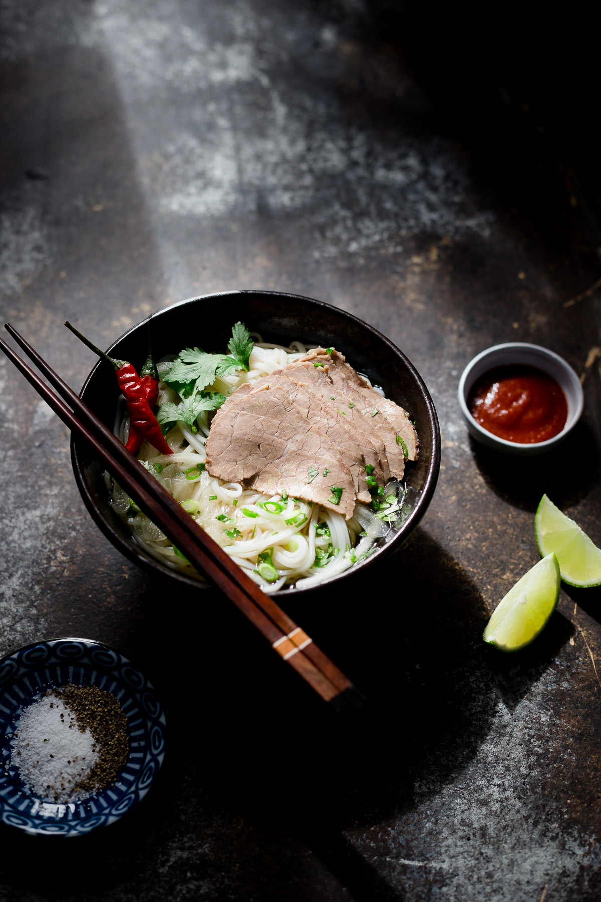A bowl of lemongrass beef and noodle soup served with some hot sauce, lime wedges, and salt and pepper.