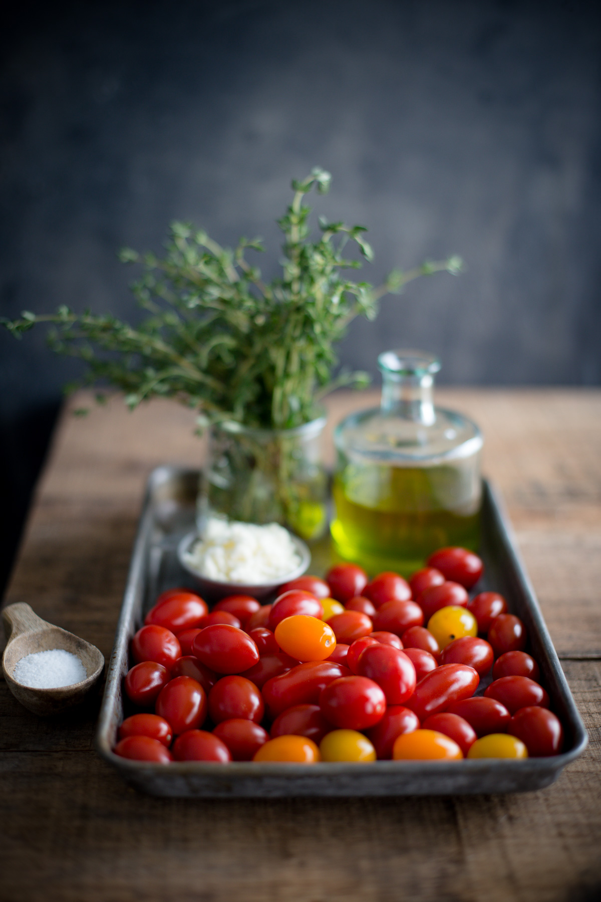 Ingredients for roasted tomatoes set out on a metal tray - tomatoes, olive oil, fresh thyme, feta, and kosher salt.