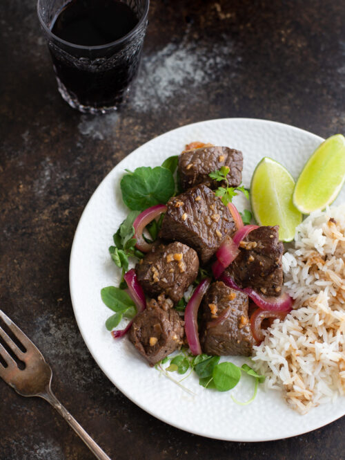 Shaking beef on a white plate next to a fork and glass of wine.