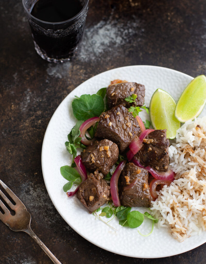 Shaking beef on a white plate next to a fork and glass of wine.