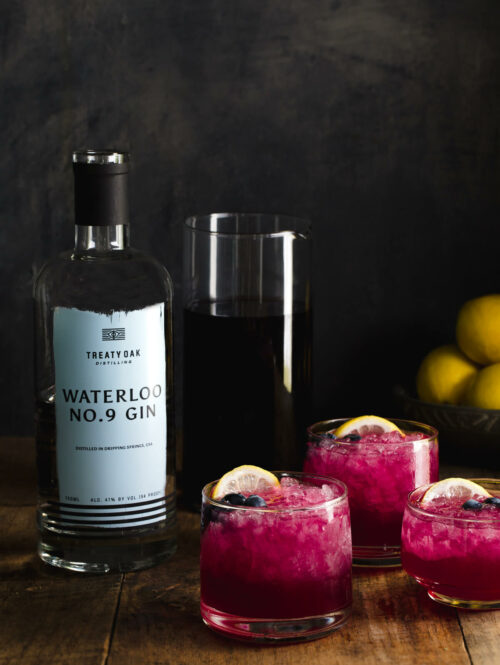 Three glasses of gin cocktail with lemon juice and blueberry-lavender-juniper simple syrup next to a bottle of gin.
