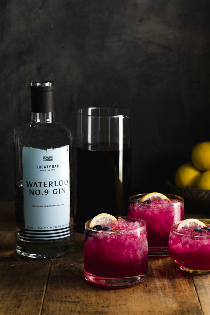 Three glasses of gin cocktail with lemon juice and blueberry-lavender-juniper simple syrup next to a bottle of gin.