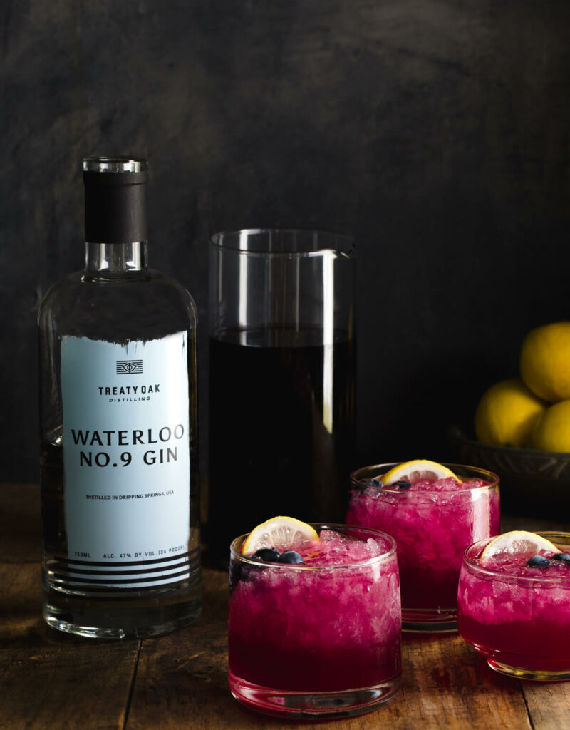 Three glasses of gin cocktail with lemon juice and blueberry-lavender-juniper simple syrup next to a bottle of gin.