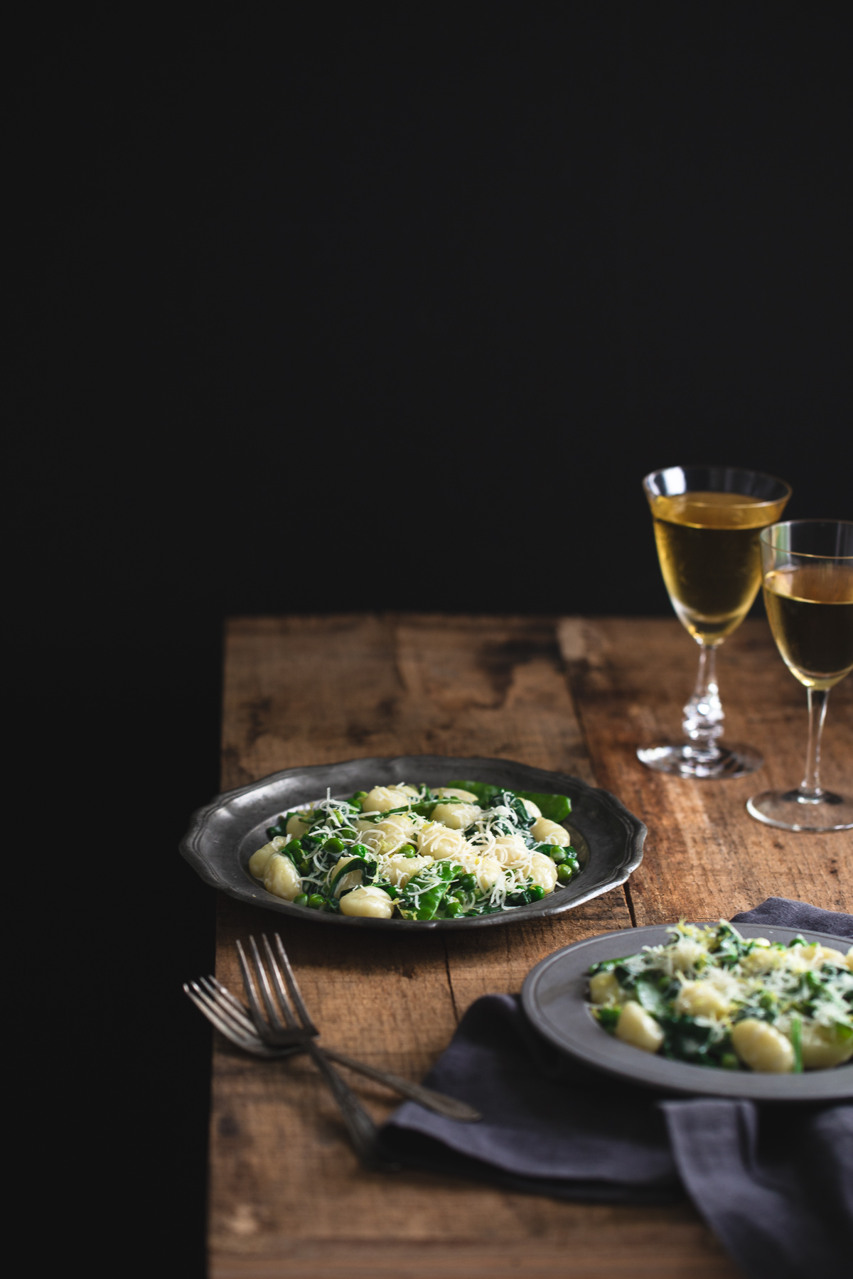 Plates of lemon gnocchi with spinach and peas on a wooden tabletop served with white wine.