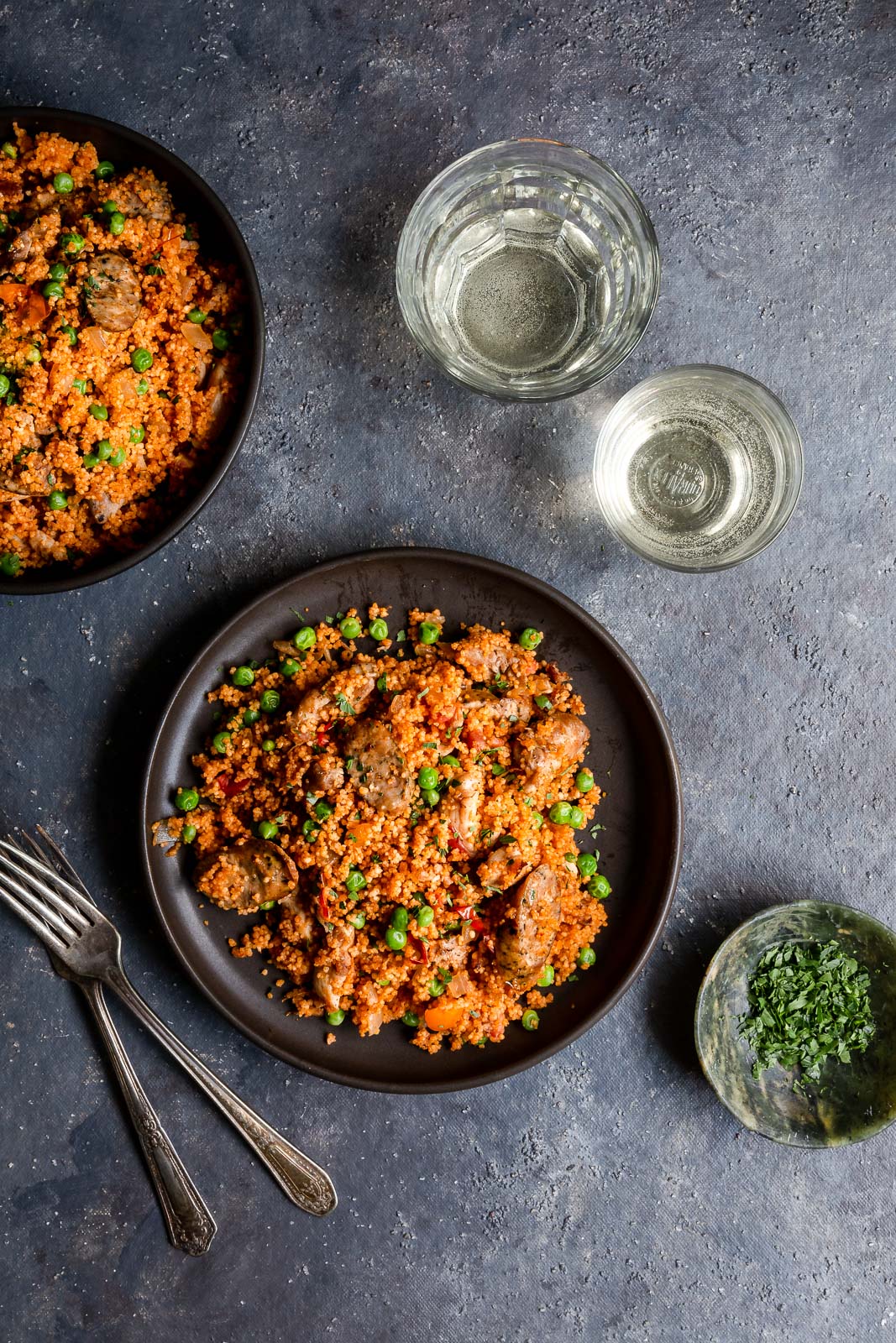 Overhead view of chicken and sausage couscous paella  on a brown plates with glasses of white wine.