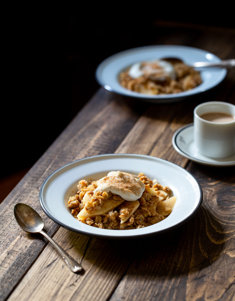 Two plates of breakfast apple crisp and a cup of coffee on a wooden table.