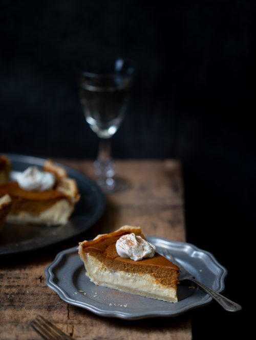 Slice of layered pumpkin cheesecake pie on pewter plate on wooden board with more pie slices and glass of white wine in the background.