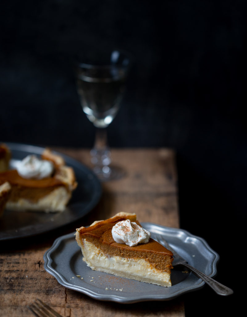 Slice of layered pumpkin cheesecake pie on pewter plate on wooden board with more pie slices and glass of white wine in the background.