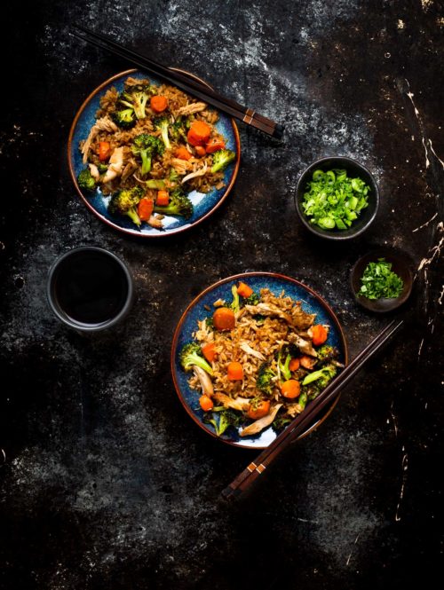 Vegetable and chicken fried rice on two blue plates with wooden chopsticks shown with a small bowl of chopped green onions, a small bowl of chopped cilantro and a cup of tea.