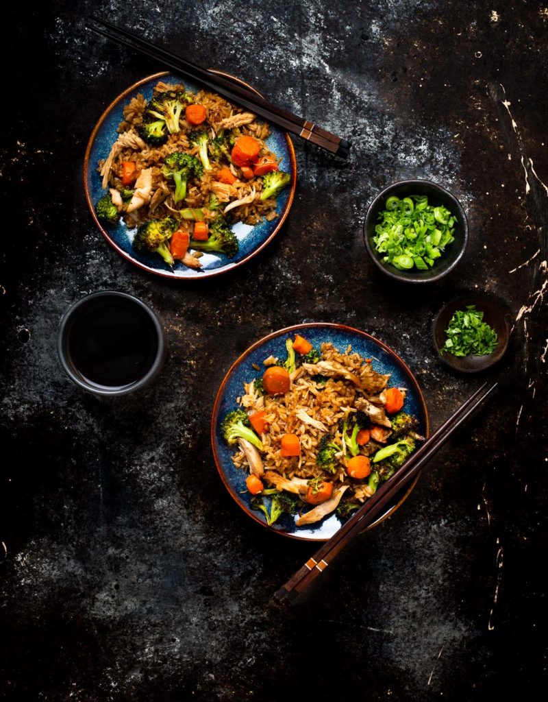 Vegetable and chicken fried rice on two blue plates with wooden chopsticks shown with a small bowl of chopped green onions, a small bowl of chopped cilantro and a cup of tea.