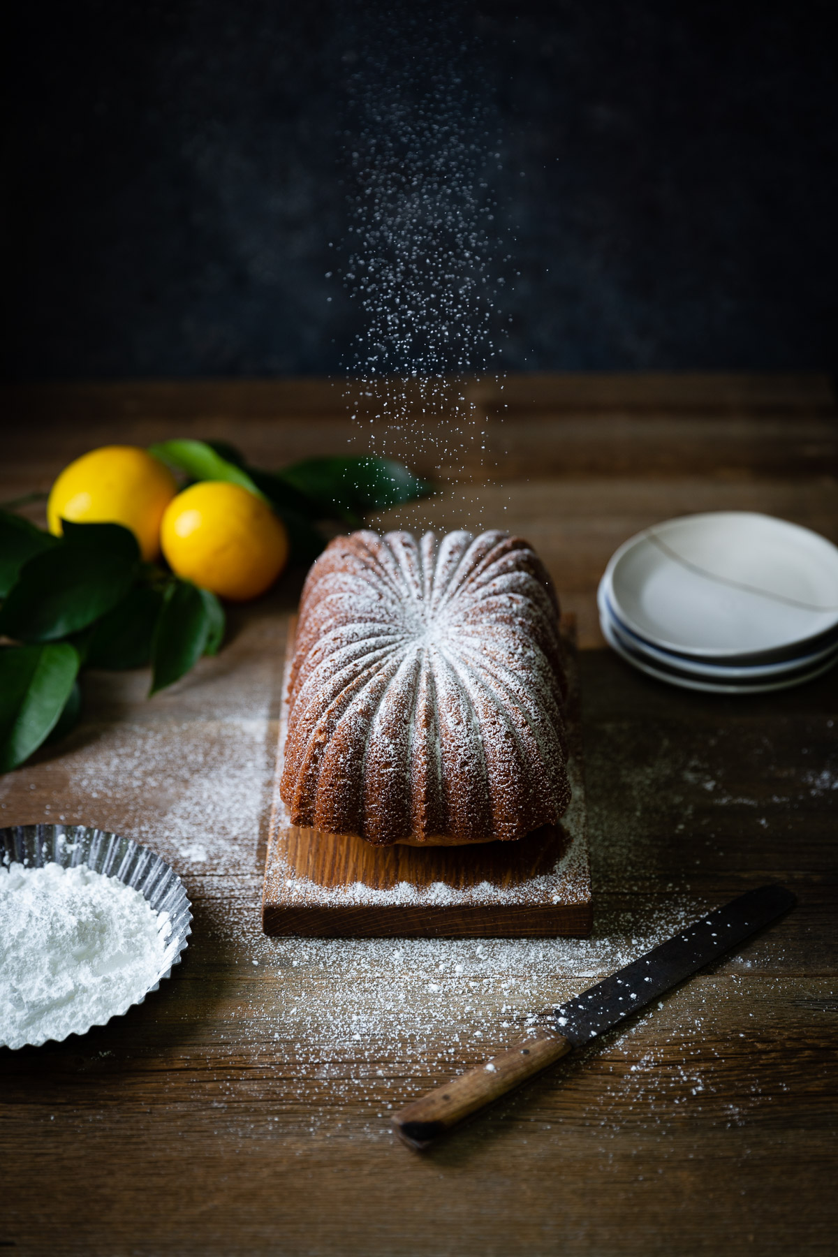 A lemon tea cake with lemon drizzle on a dark brown table being sprinkled with powdered sugar. Two lemons, a stack of dessert plates, and a dish of powdered sugar are set around the cake.