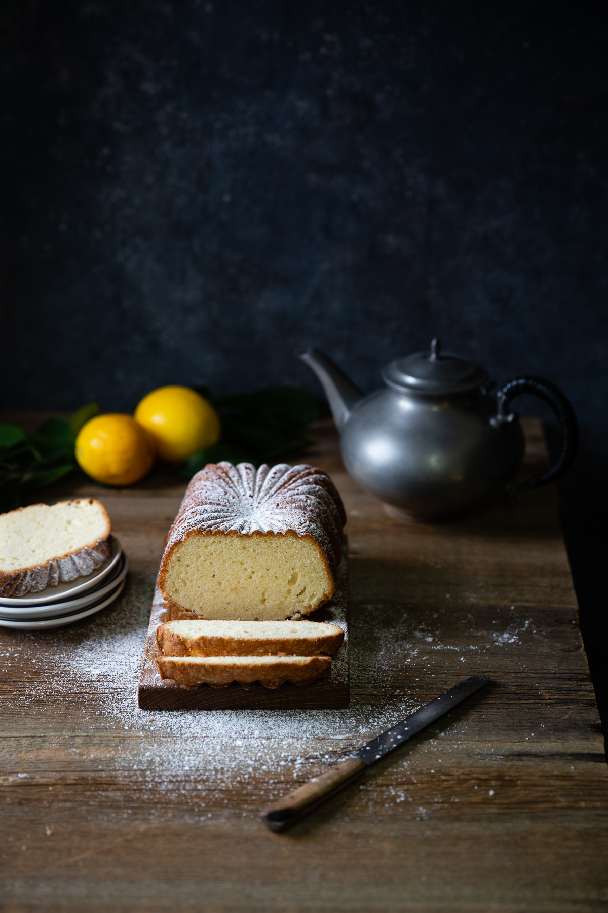 A lemon tea cake loaf with two slices cut and sprinkled with powdered sugar on a brown table top. Two lemons, a slice of cake on a stack dessert plates, and a pewter teapot are set around the cake.