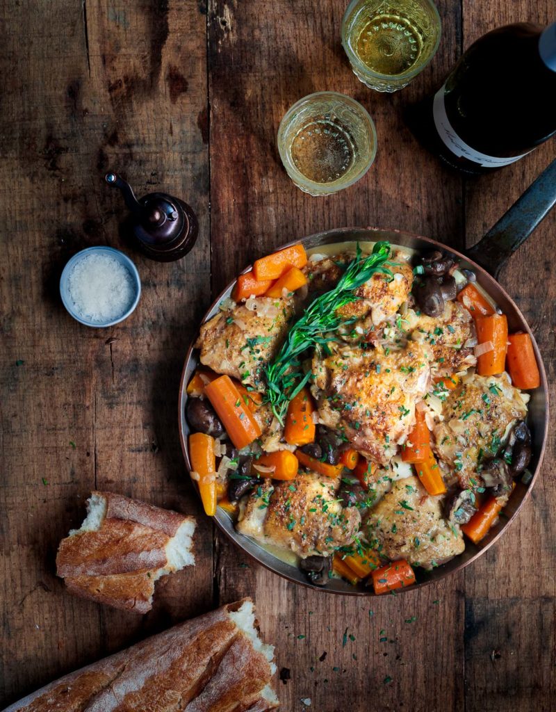 Braised chicken and carrots in a pan on a wooden surface with white wine and a baguette.