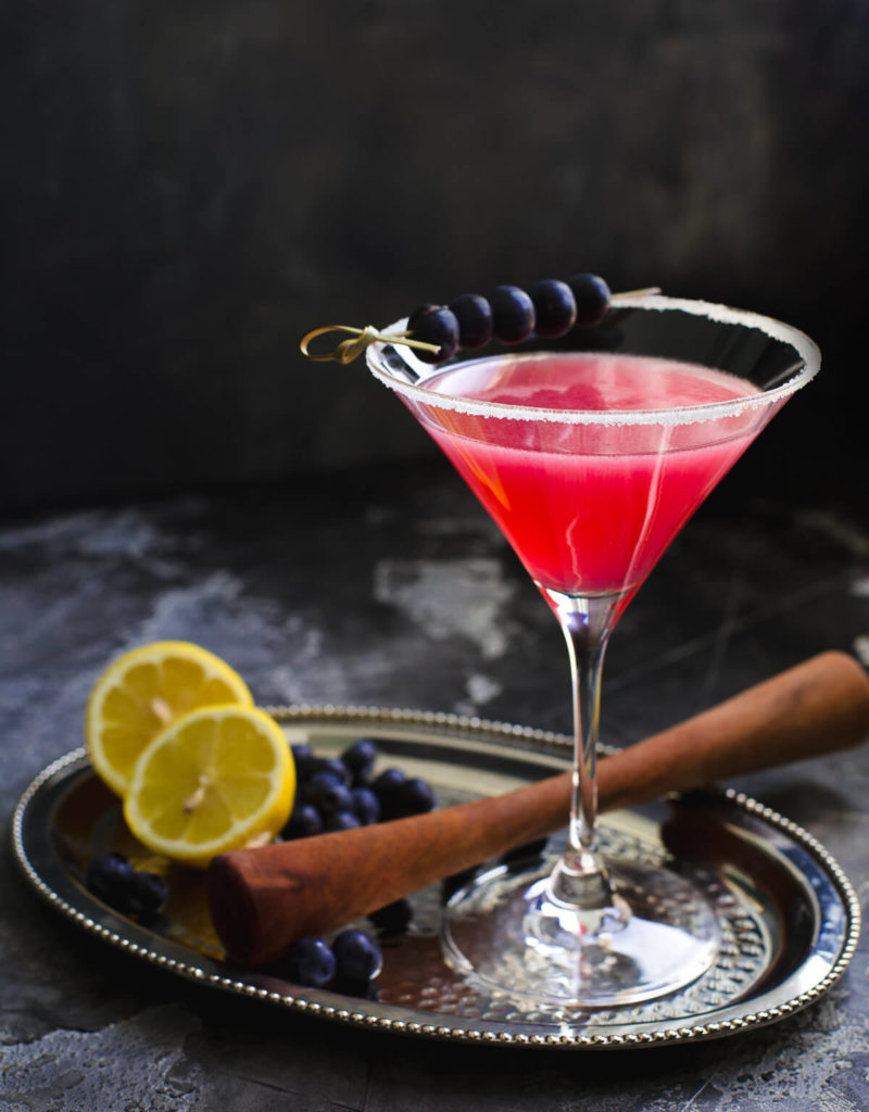 Blueberry Lemon Drop cocktail in a tall martini glass garnished with blueberries on a metal tray.