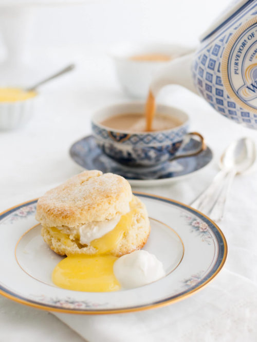 Cream scone, lemon curd, and whipped cream on a china plate with a cup of tea being poured in the background.