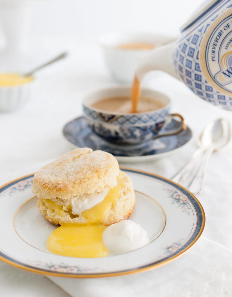 Cream scone, lemon curd, and whipped cream on a china plate with a cup of tea being poured in the background.