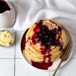 Overhead image of a stack of lemon ricotta pancakes topped with blueberry compote on a white plate on a white tile tabletop pictured with a small bowl of lemon butter and a container of blueberry compote.