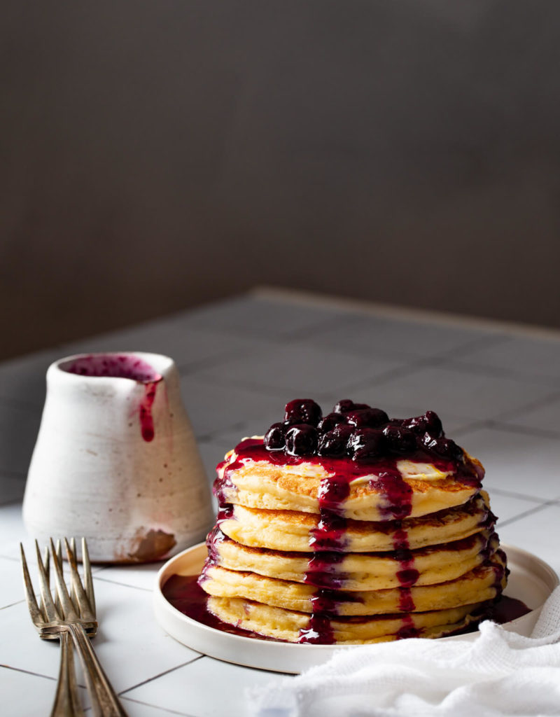 Stack of lemon ricotta pancakes topped with blueberry compote on a white plate on a white tile tabletop.