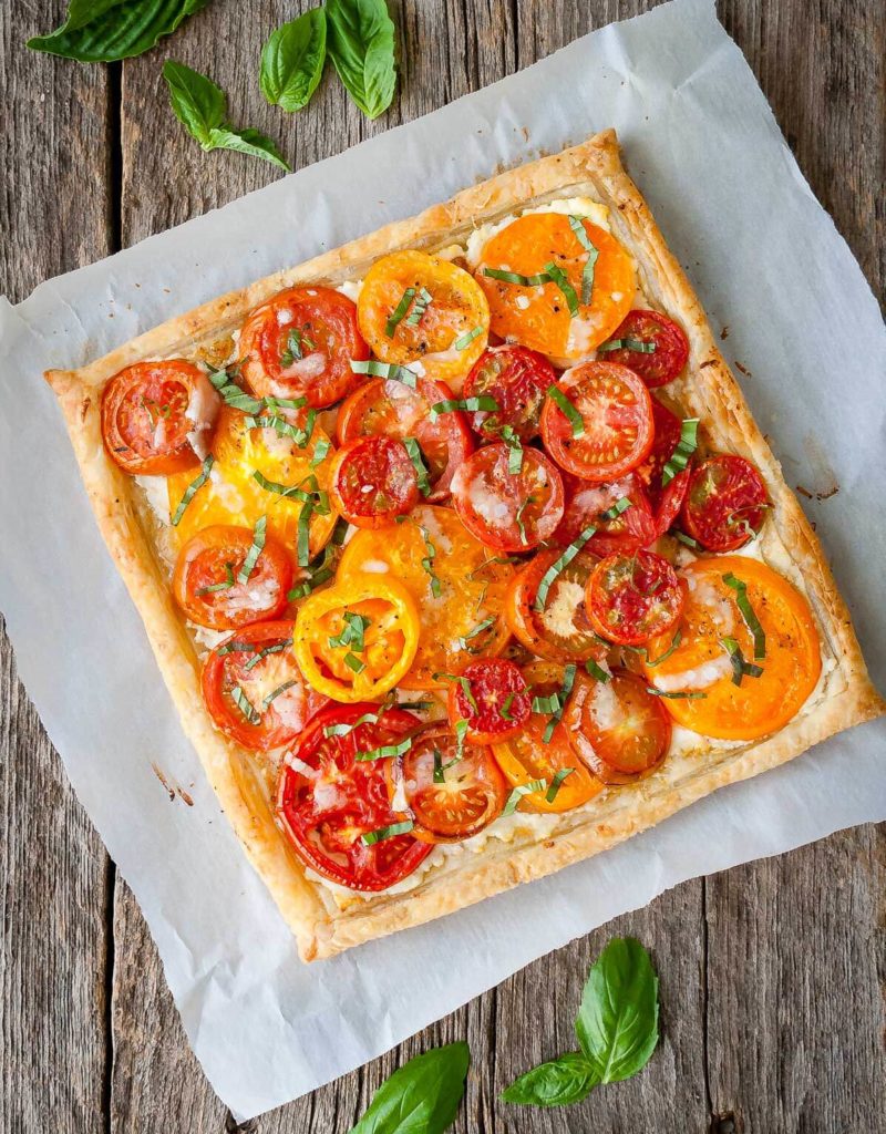 Tomato tart with goat cheese and caramelized onions on a piece of parchment paper with loose basil leaves.