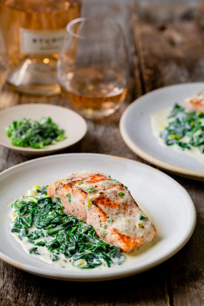 A plate of pan seared salmon with creamed spinach on the side shown with a bottle of Rosé wine and a wine glass.