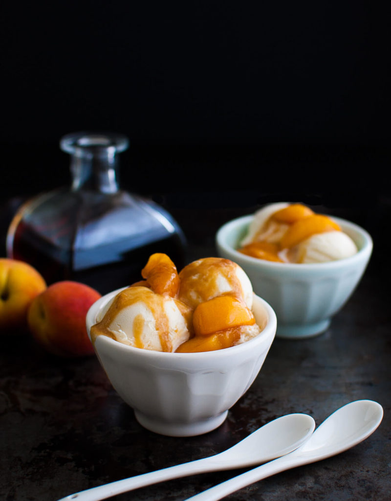 Two bowls of ice-cream topped with peaches foster shown with two white spoons and some peaches and a bottle of rum in the background.