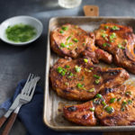 Sheet pan of pork chops with blue napkin, fork, and knife in the front and dish of scallion oil and glasses of white wine in the background.