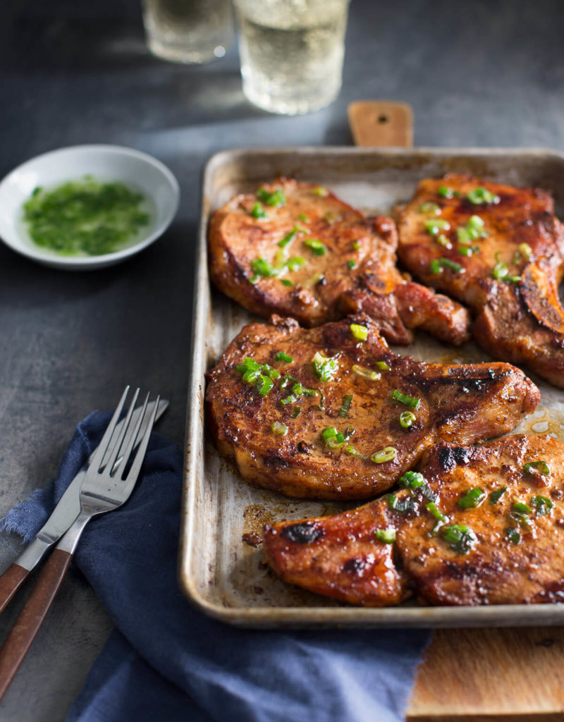 Sheet pan of pork chops with blue napkin, fork, and knife in the front and dish of scallion oil and glasses of white wine in the background.