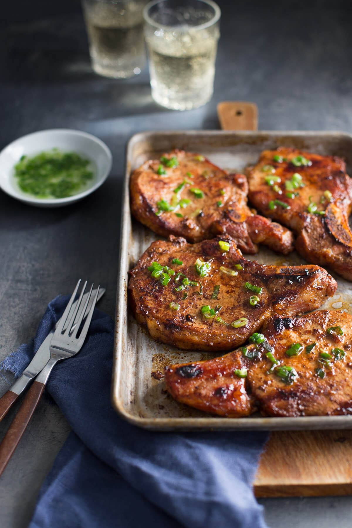 Sheet pan of pork chops with blue napkin, fork, and knife in the front and dish of scallion oil and glasses of white wine in the background.