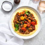 Overhead view of cherry tomato ragout and polenta in a serving dish next to some glasses of rosé.