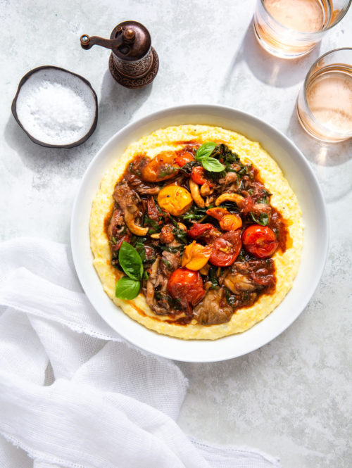 Overhead view of cherry tomato ragout and polenta in a serving dish next to some glasses of rosé.