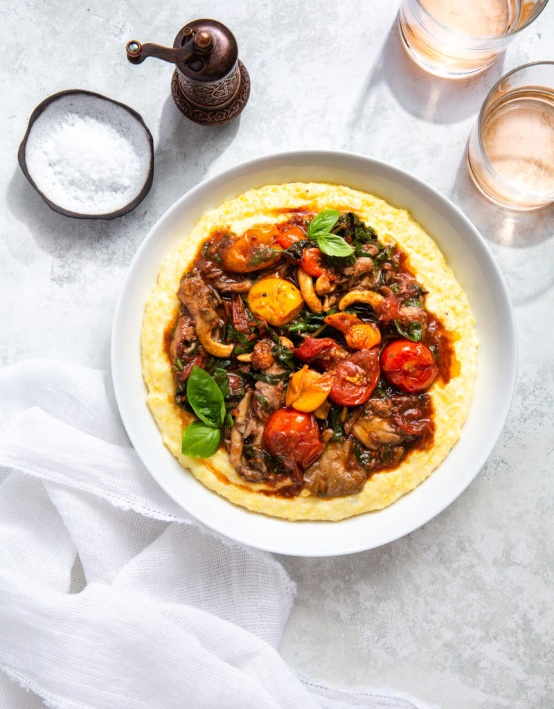 Overhead view of cherry tomato ragout and polenta in a serving dish next to some glasses of rosé.